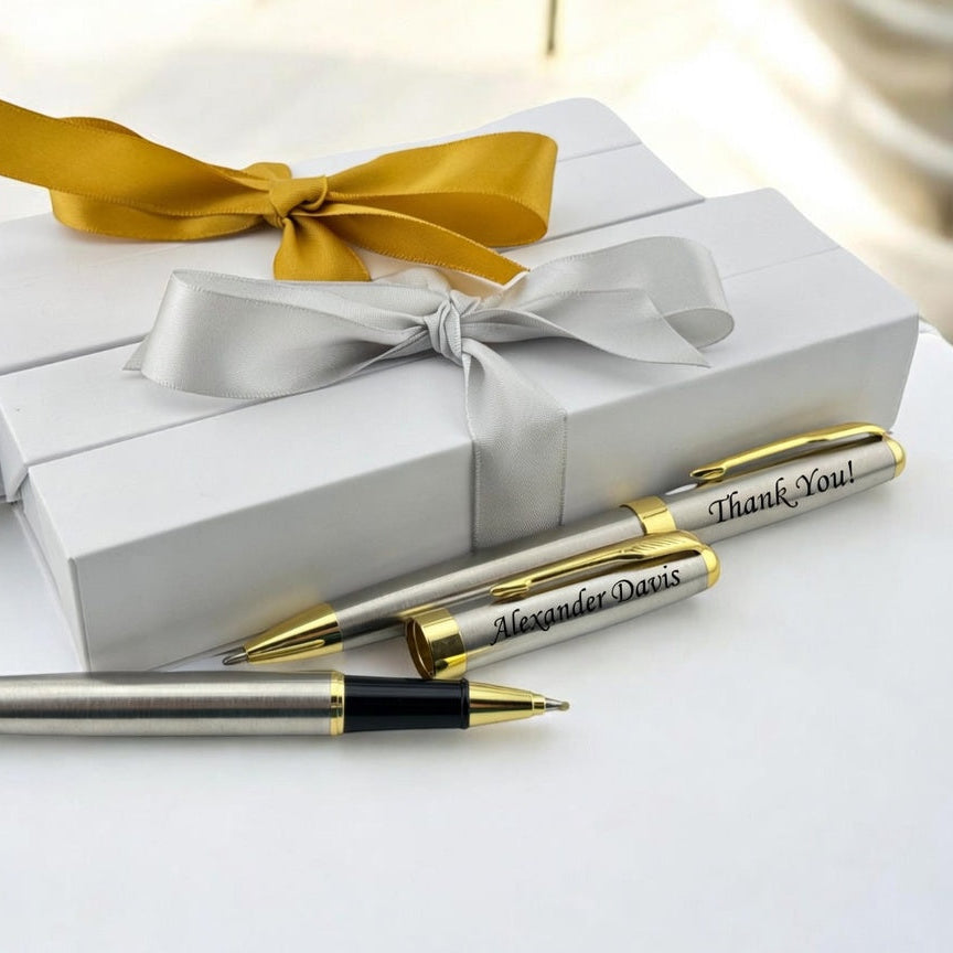 Three personalized metal pens with gold accents are arranged in front of two white gift boxes tied with satin ribbons, one gold and one silver, on a white surface.