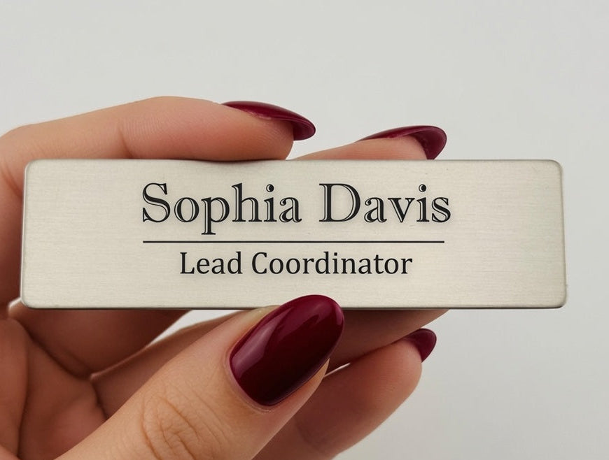 A rectangular brushed-metal nameplate with engraved black text reading 'Sophia Davis' and 'Lead Coordinator', held by a hand with dark red manicured nails against a plain light background. The text 'Style 3' appears in the lower right corner.