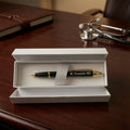 A personalized black pen with gold trim and 'M. Fernandez MD' engraved on the clip, presented in an open white gift box on a dark wooden desk with a stethoscope in the background.