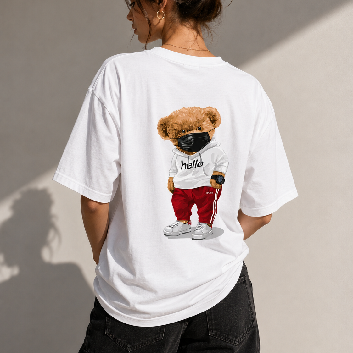 A woman shown from behind wearing a white oversized graphic T-shirt. The shirt features a cartoon teddy bear wearing a black face mask, white hoodie with 'hello' text, red pants, and white sneakers.