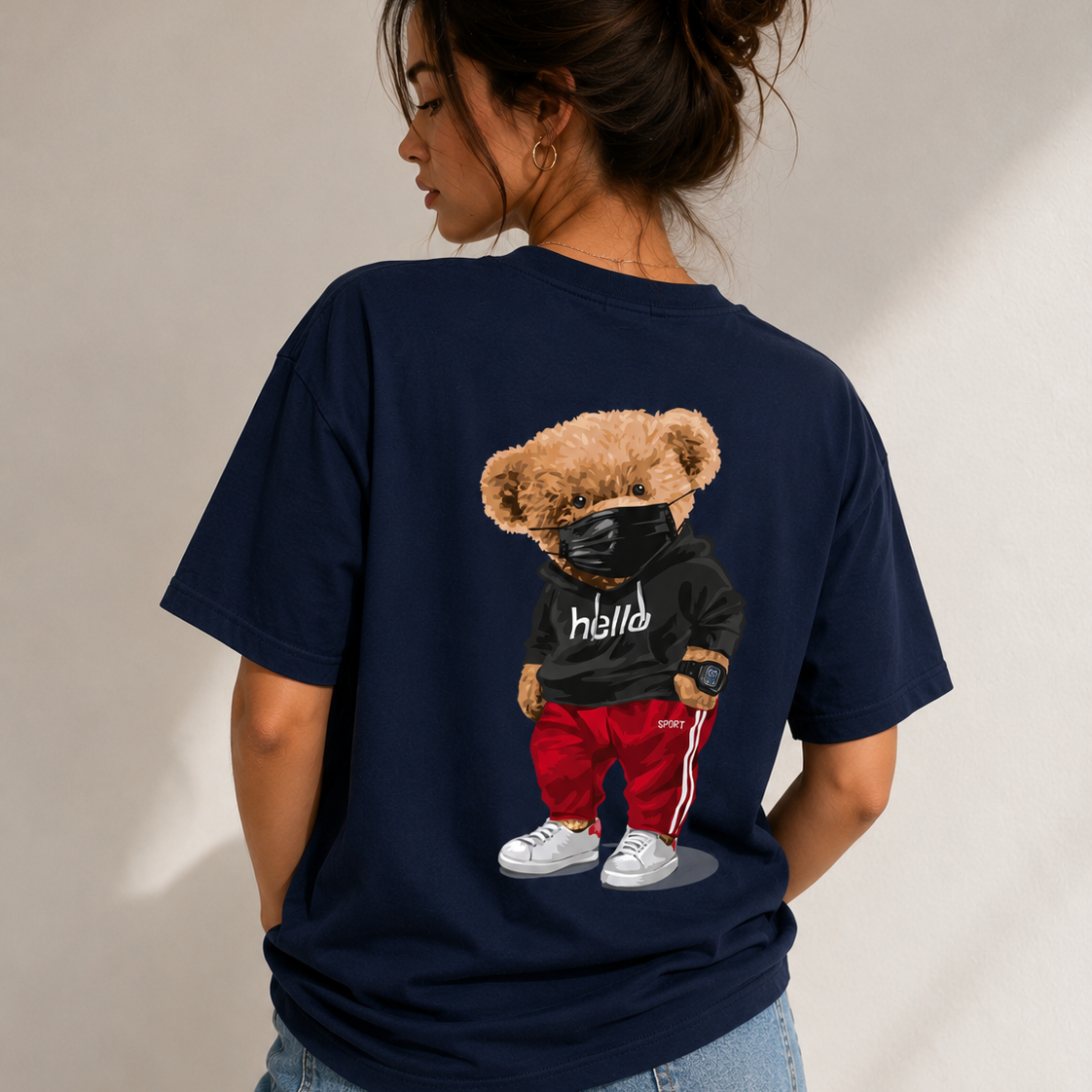 A woman shown from the back wearing a navy blue oversized t-shirt with a large graphic of a teddy bear wearing a black hoodie, red pants, and white sneakers. She is also wearing light wash blue jeans against a neutral beige background.
