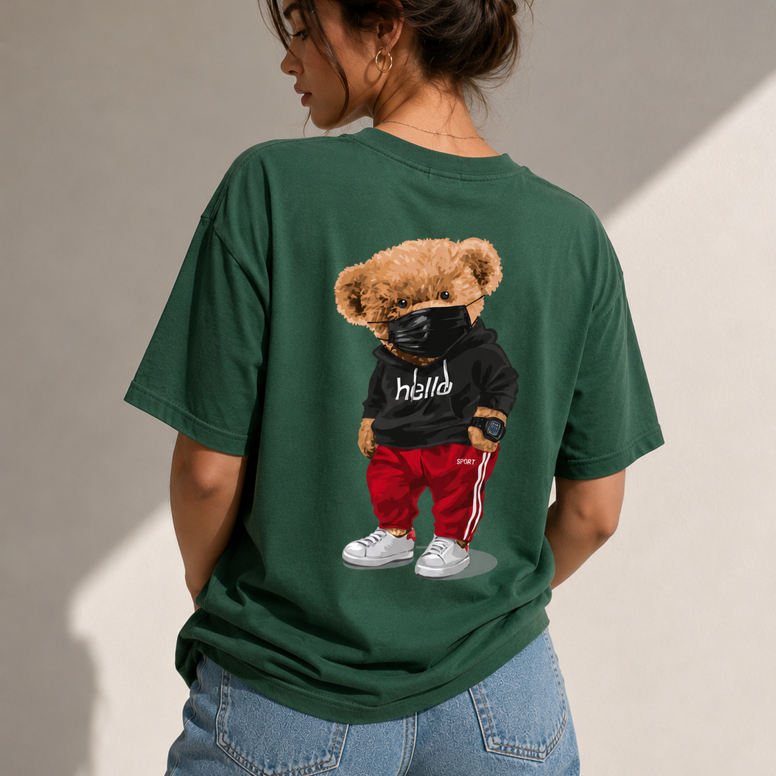 A woman shown from the back wearing a dark green oversized t-shirt with a large graphic of a teddy bear wearing a black hoodie, red pants, and white sneakers. She is also wearing blue jeans against a neutral beige wall with soft diagonal shadows.