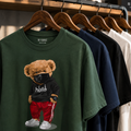 A green graphic t-shirt featuring a cartoon teddy bear wearing a black hoodie and red pants, hanging on a wooden rack alongside other t-shirts in navy, black, white, and beige. The brand 'BE VISIBLE COLLECTION' is printed inside the collar.