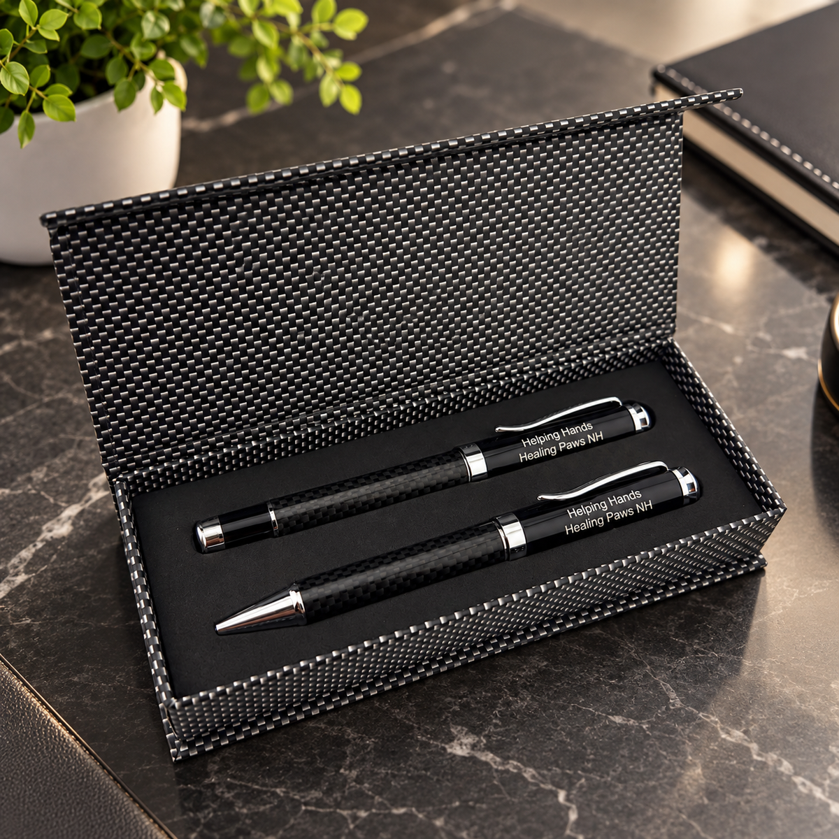 A personalized black carbon-fiber-style pen set presented in an open textured black gift box on a dark marble desk, with a potted plant and notebook in the background.