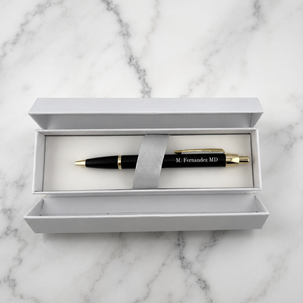 A black ballpoint pen with gold trim and 'M. Fernandez MD' printed on the barrel, presented in an open white gift box on a white marble surface.