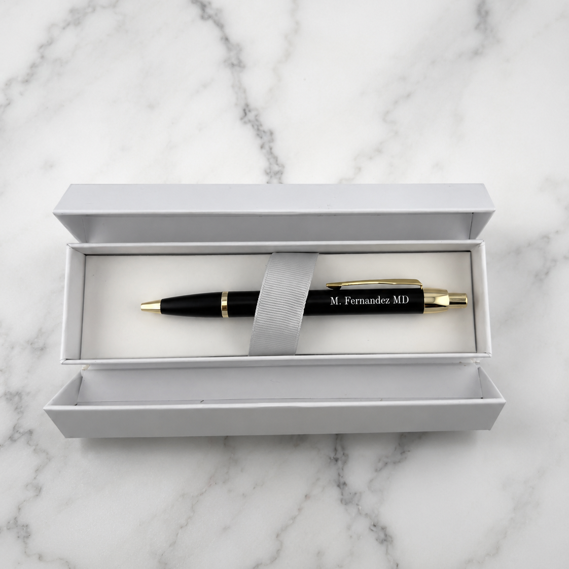 A black ballpoint pen with gold trim and 'M. Fernandez MD' printed on the barrel, presented in an open white gift box on a white marble surface.