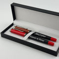 A personalized black and red pen set with a brown barrel pen, presented inside an open black gift box with a white interior. The black pen features the engraved text 'Alex Carter Forever in Focus' wrapped around a white ribbon.