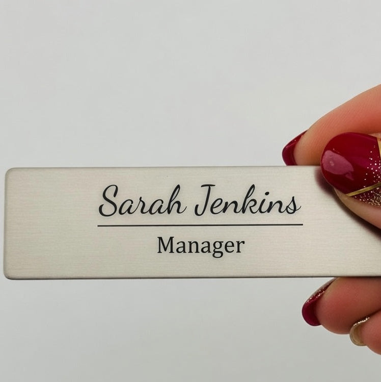 A rectangular brushed-metal name badge holder engraved with 'Sarah Jenkins Manager', held by a hand with red and gold nail polish against a plain light background. The text 'Style 1' appears in the bottom right corner.