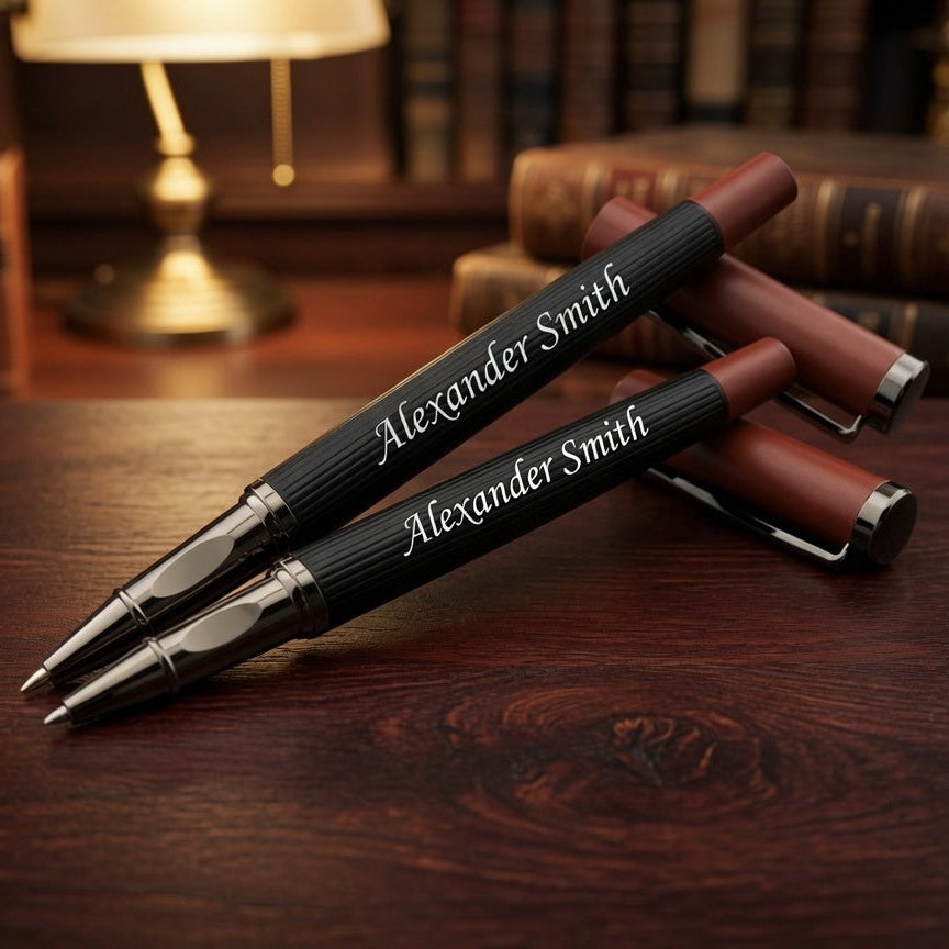 Three personalized ballpoint pens with black ribbed barrels, silver metal tips, and reddish-brown accents, resting on a dark wooden desk with books and a lamp in the background. The pens are engraved with the name 'Alexander Smith'.