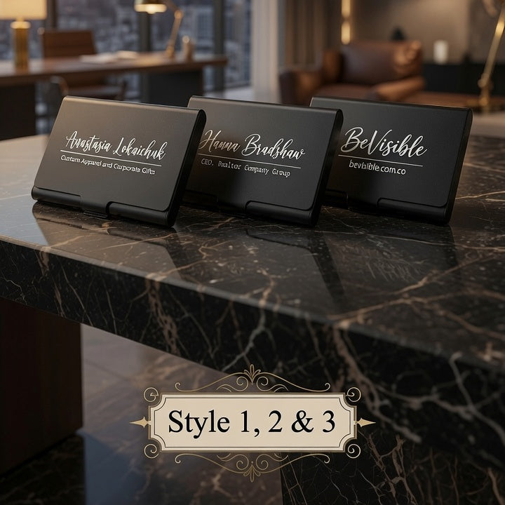 Three black rectangular name card stands or desk signs with elegant white script text are arranged on a dark marble tabletop in a modern room. Large windows behind show a city skyline at sunset, and warm interior lighting is visible.