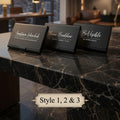Three black rectangular name card stands or desk signs with elegant white script text are arranged on a dark marble tabletop in a modern room. Large windows behind show a city skyline at sunset, and warm interior lighting is visible.