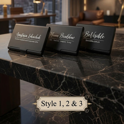 Three black rectangular name card stands or desk signs with elegant white script text are arranged on a dark marble tabletop in a modern room. Large windows behind show a city skyline at sunset, and warm interior lighting is visible.