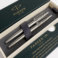 Three Parker ballpoint pens in a sleek silver finish, presented inside an open cream-colored gift box with dark green branded packaging. The pens are secured by a dark navy elastic band, and the brand name 'PARKER' is clearly visible on both the pen barrels and the box lid.