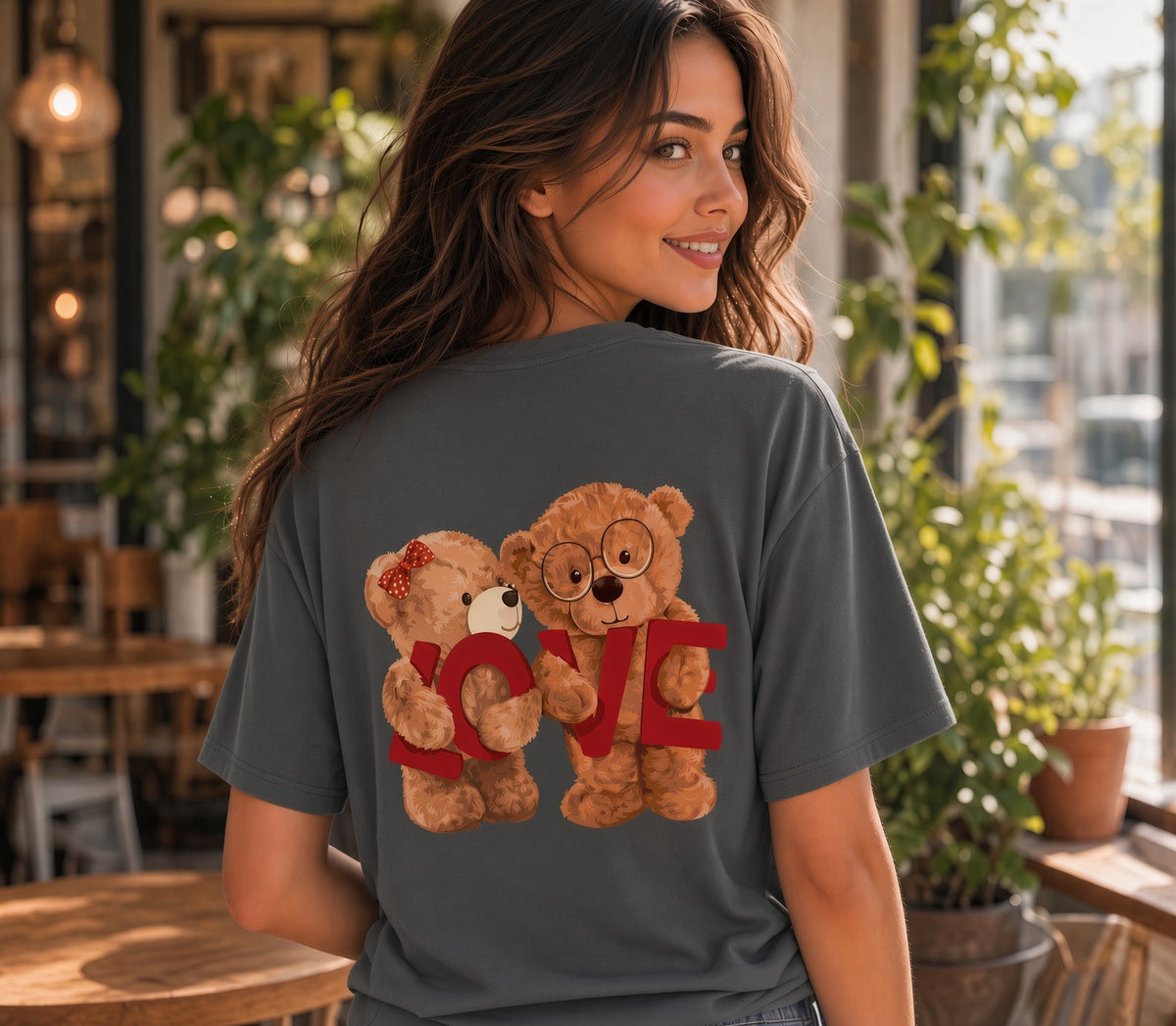 A woman wearing a dark gray t-shirt with a graphic of two teddy bears holding a red 'LOVE' sign, photographed in a cozy cafe setting.