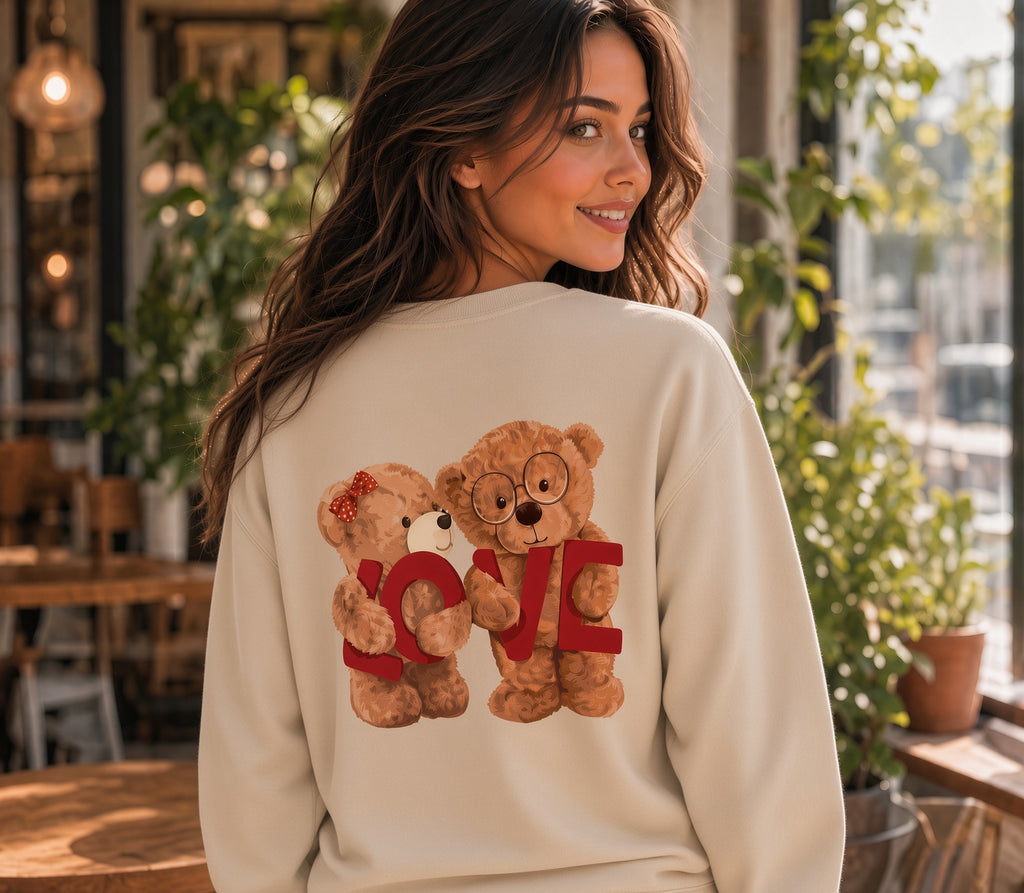 A woman wearing a cream-colored sweatshirt with a graphic of two teddy bears holding a red 'LOVE' sign, photographed in a warm, sunlit cafe setting.