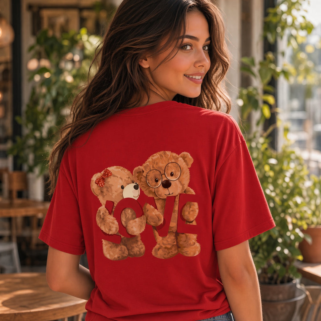 A woman wearing a red short-sleeve t-shirt with a graphic of two teddy bears holding a heart-shaped 'LOVE' sign, photographed in a warm, sunlit cafe setting.