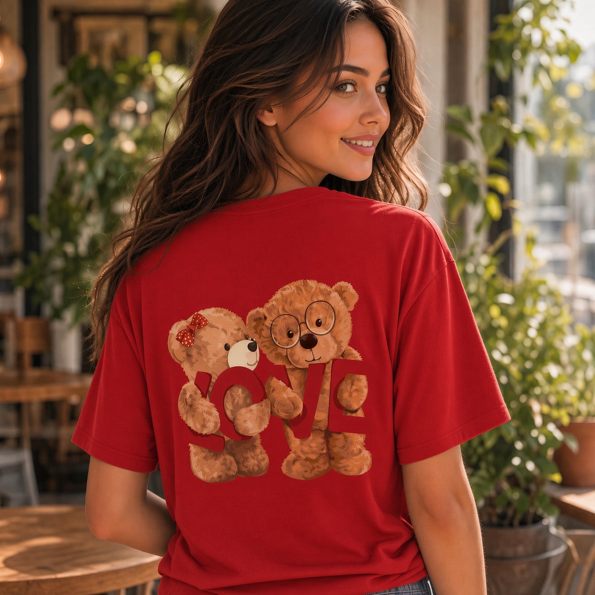 A woman wearing a red short-sleeve t-shirt with a graphic of two teddy bears holding a heart-shaped 'LOVE' sign, photographed in a warm, sunlit cafe setting.