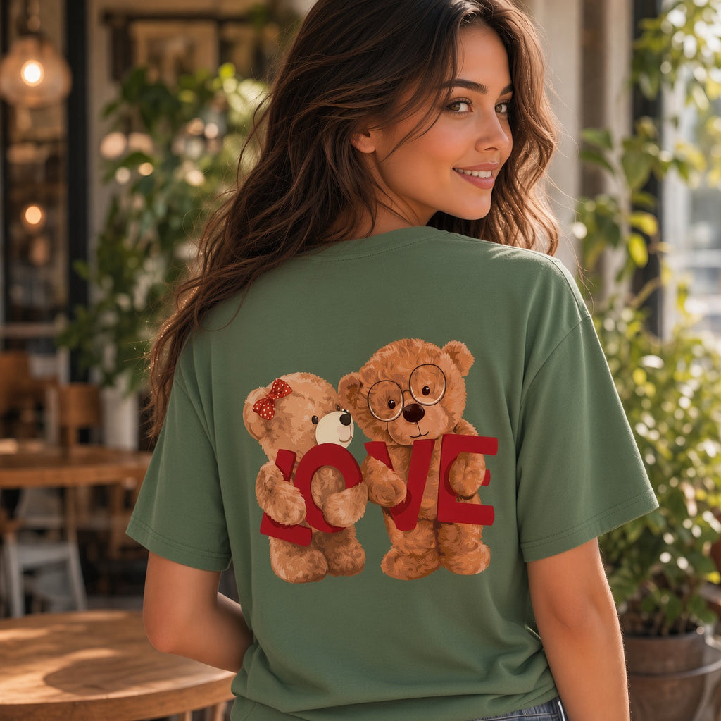 A woman wearing an olive green t-shirt with a graphic of two teddy bears holding a red 'LOVE' sign, photographed in a cozy, sunlit cafe setting.