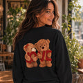 A woman wearing a black sweatshirt with a graphic of two teddy bears holding a red 'LOVE' sign, photographed in a cozy indoor cafe setting.