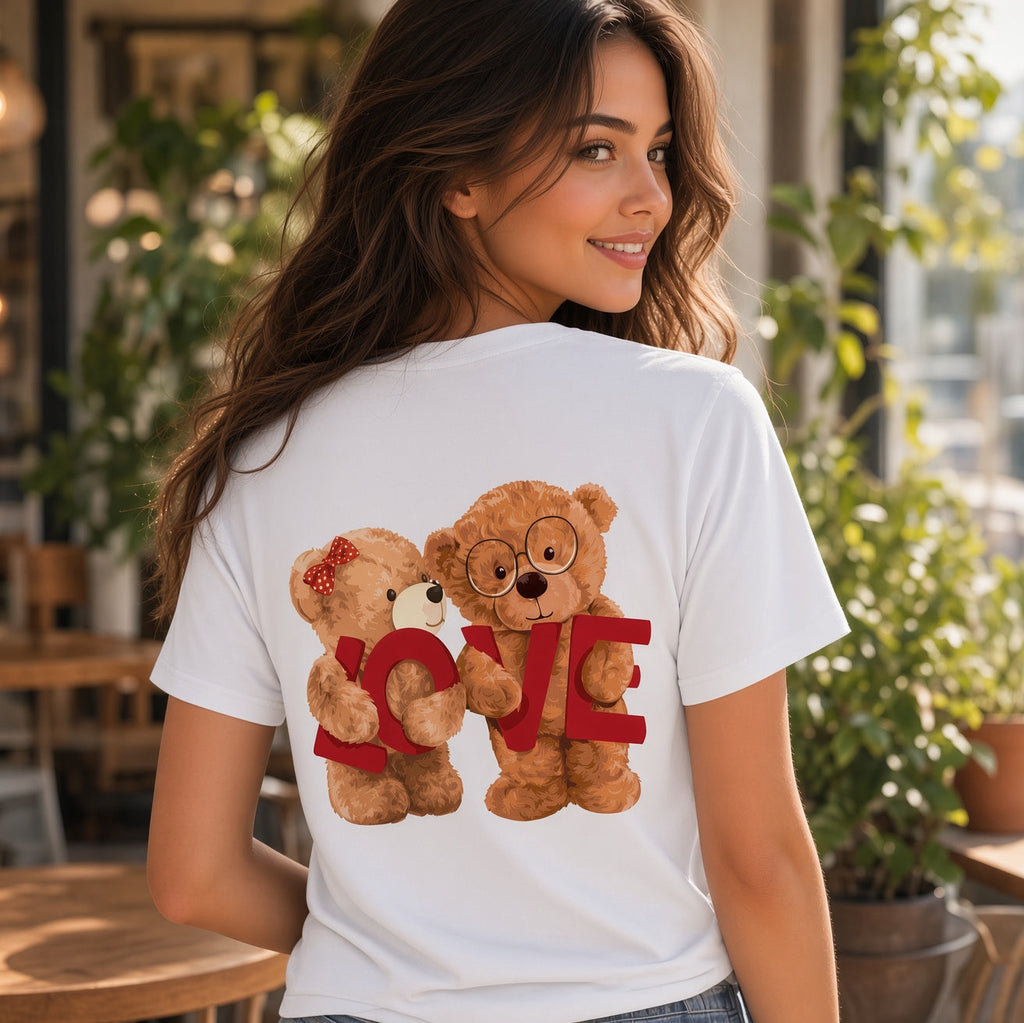 A woman wearing a white short-sleeve T-shirt with a graphic of two teddy bears hugging behind a red 'LOVE' text design, photographed in a bright indoor cafe setting.