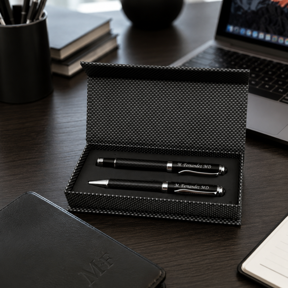 A black textured pen and matching pen case set displayed open on a dark wood desk, with a laptop, notebook, and small plant in the background.