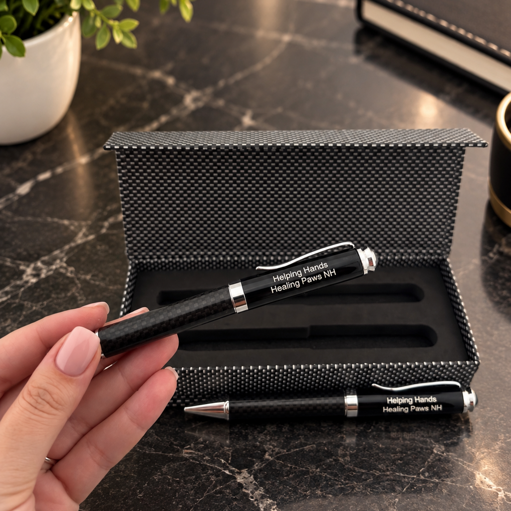 A hand holds a black pen with a carbon fiber pattern and silver accents above an open matching black textured gift box containing two personalized pens on a dark marble tabletop.