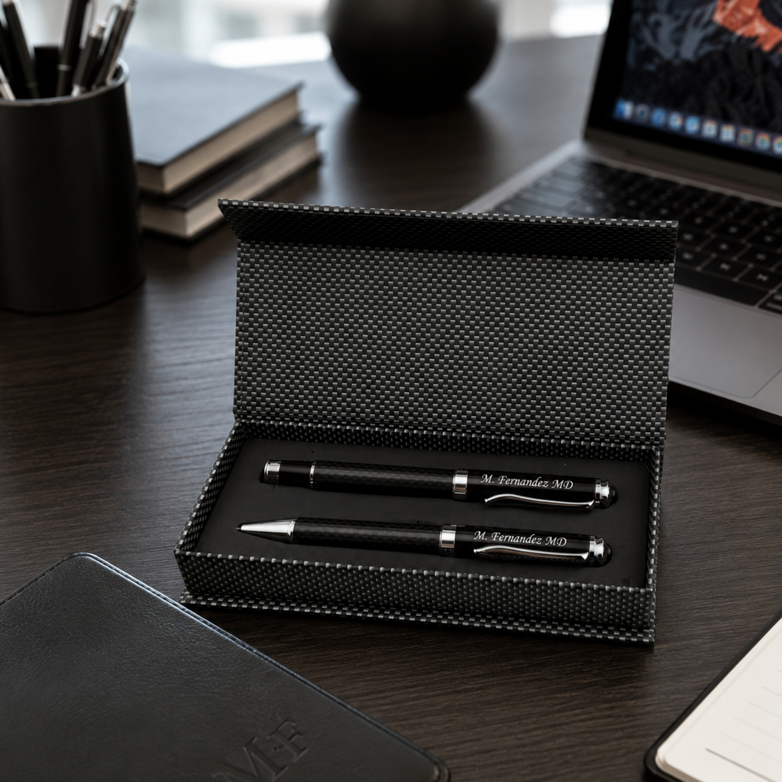 A black textured pen and matching pen case set displayed open on a dark wood desk, with a laptop, notebook, and small plant in the background.