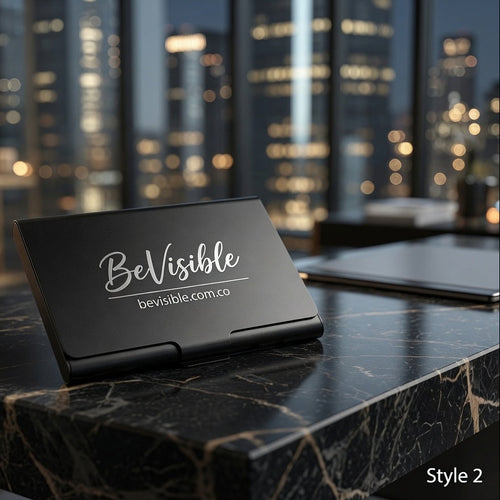 A black metal business card holder standing on a dark marble desk, printed with the white text 'BeVisible' and 'bevisible.com.