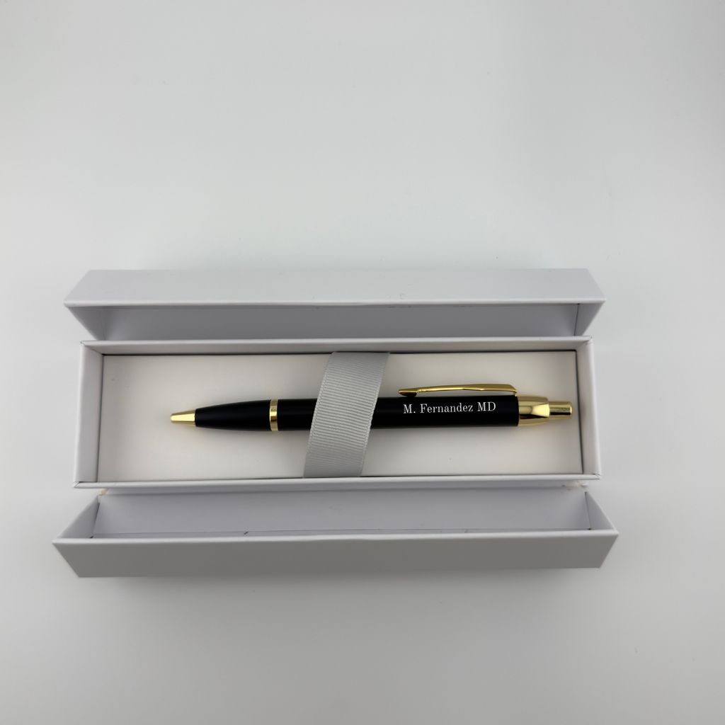 A black and gold personalized ballpoint pen with 'M. Fernandez MD' printed on the barrel, presented inside an open light gray gift box with a gray ribbon.
