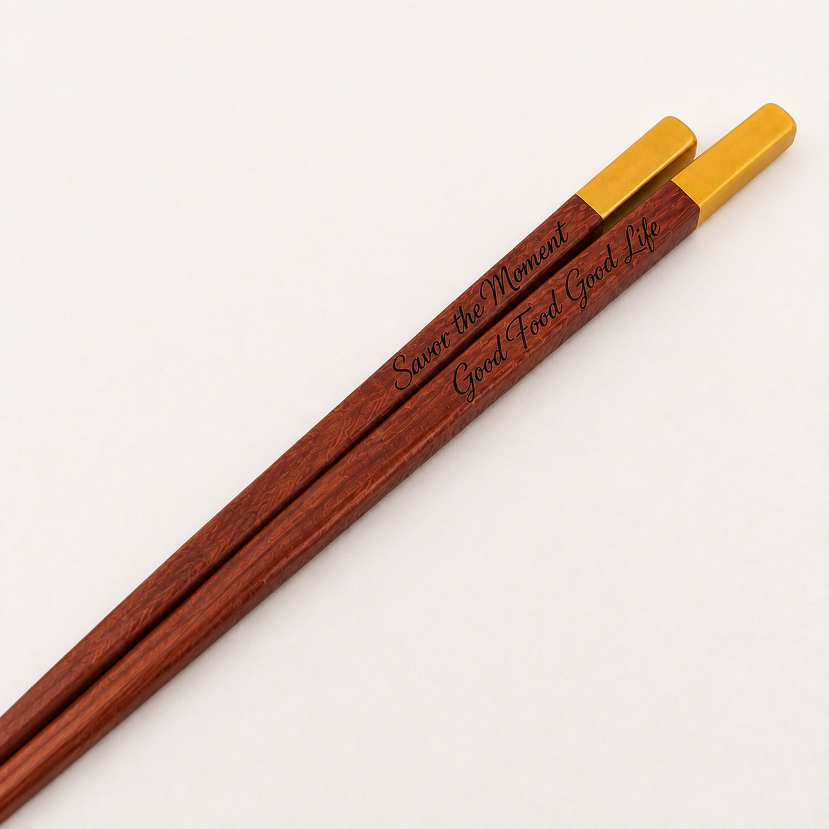 A pair of wooden chopsticks with gold metal tips, featuring engraved text on the shafts, displayed diagonally on a clean white background.