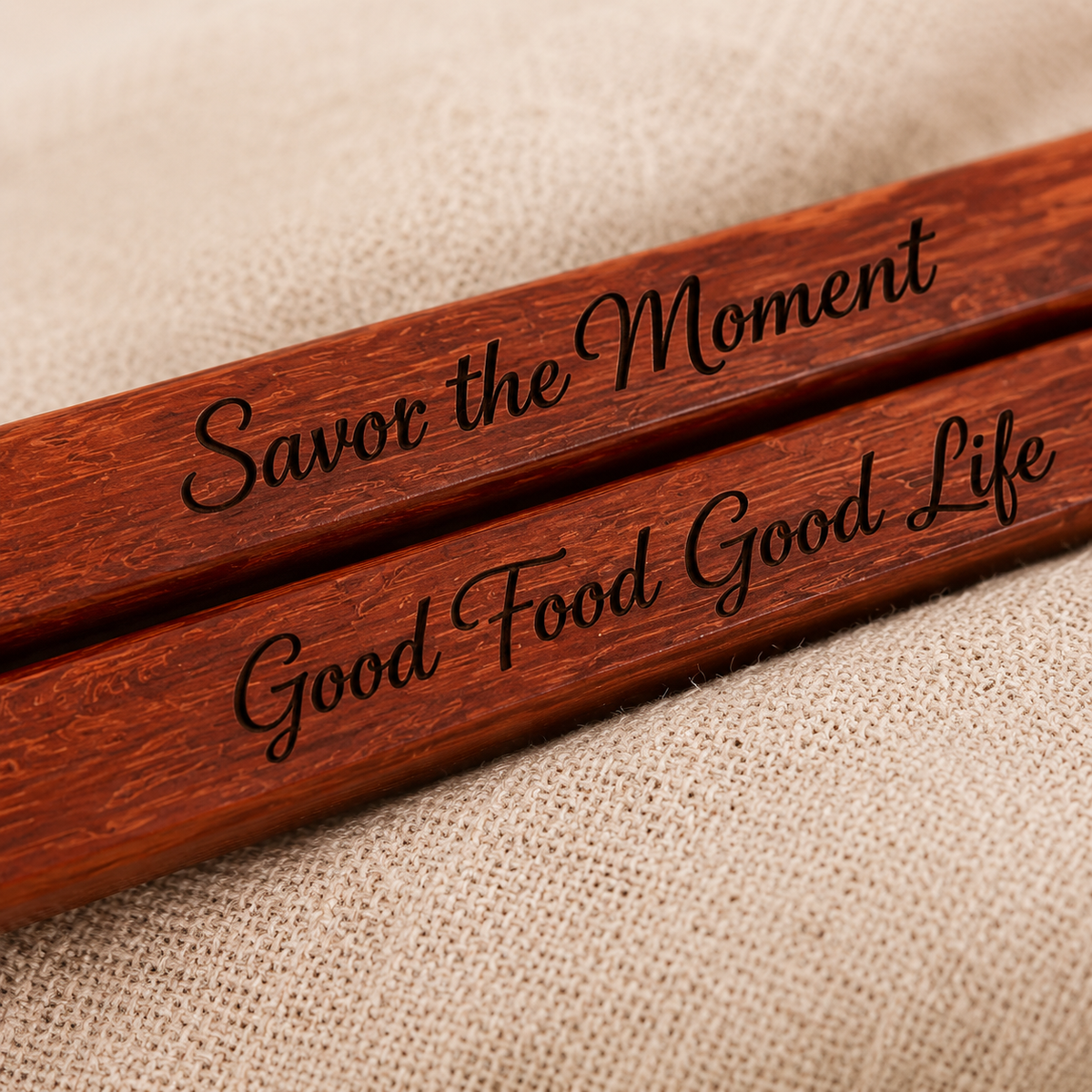 Two engraved wooden chopsticks resting on a textured beige fabric background. The chopsticks feature the phrases 'Savor the Moment' and 'Good Food Good Life' in black cursive script.
