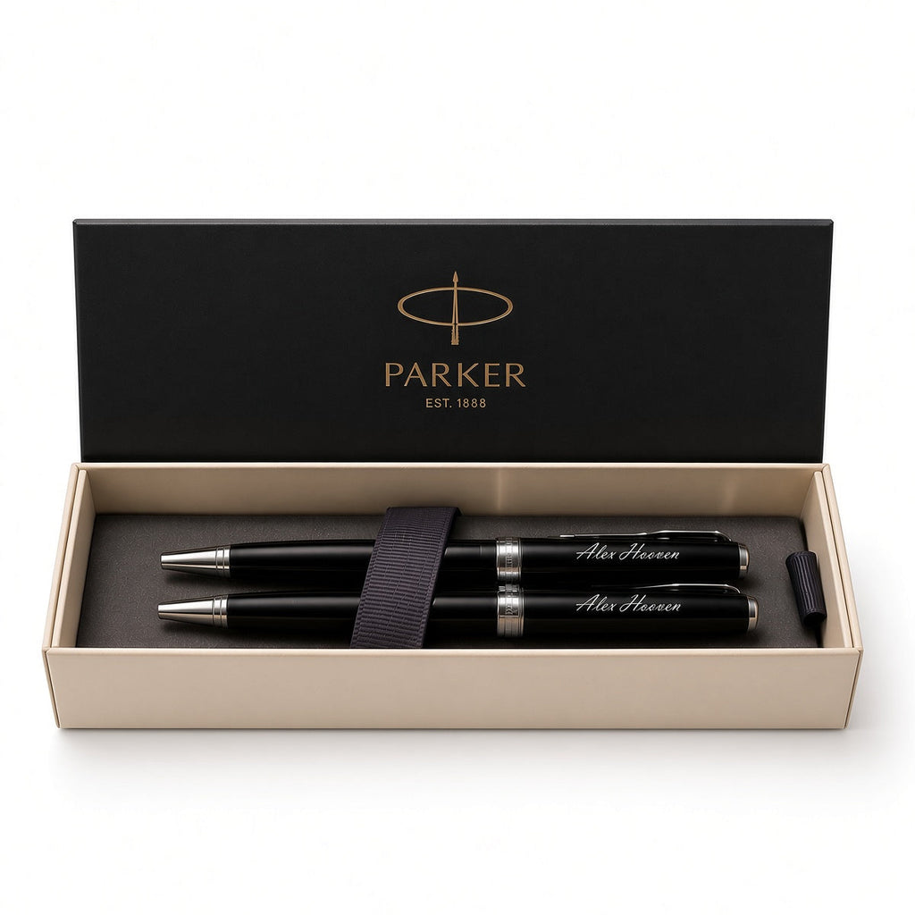 A Parker pen set in an open presentation box. The set includes two black pens with silver trim and white script engraving, a black elastic band, and a black cap, all inside a beige box with a black lid printed with the gold Parker logo.