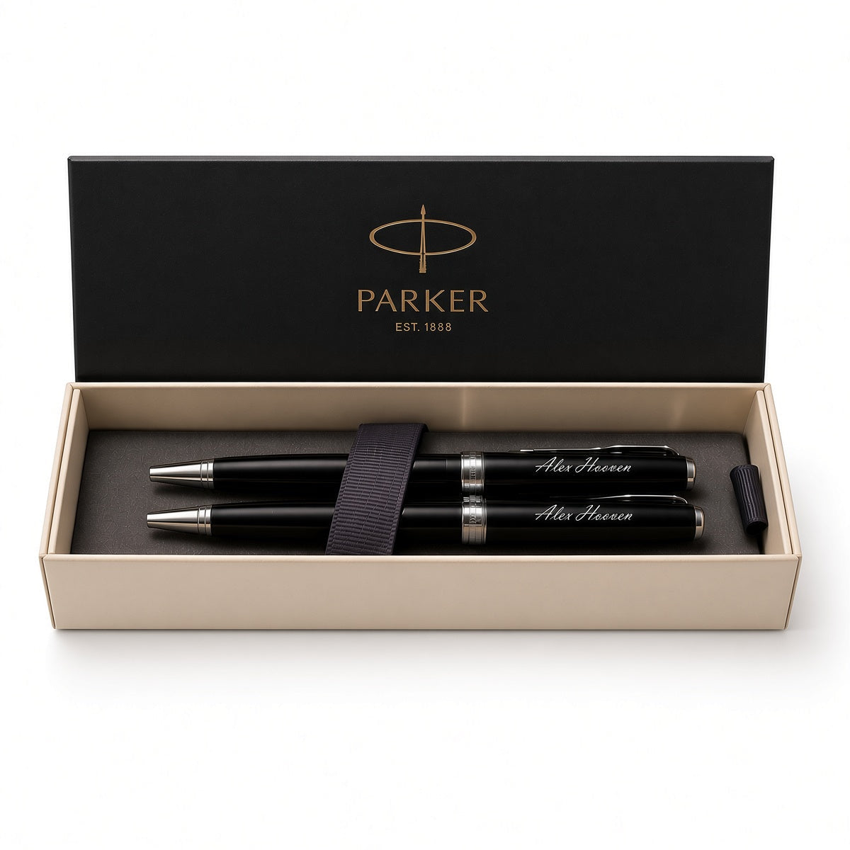 A Parker pen set in an open presentation box. The set includes two black pens with silver trim and white script engraving, a black elastic band, and a black cap, all inside a beige box with a black lid printed with the gold Parker logo.