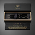 Parker IM pen set in an open black presentation box with gold trim, shown on a dark gray background. The set includes three black pens with silver and gold accents, a black ribbon, and branded packaging.