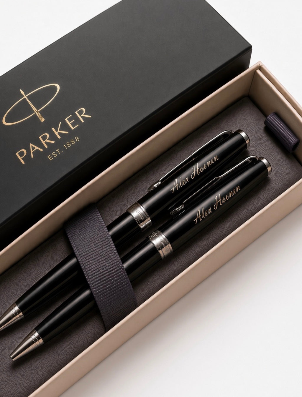 A Parker pen and ballpoint set in an open presentation box with the branded black lid visible. The set includes three black pens with silver trim and gold script personalization, plus a dark elastic band, photographed on a clean white background.