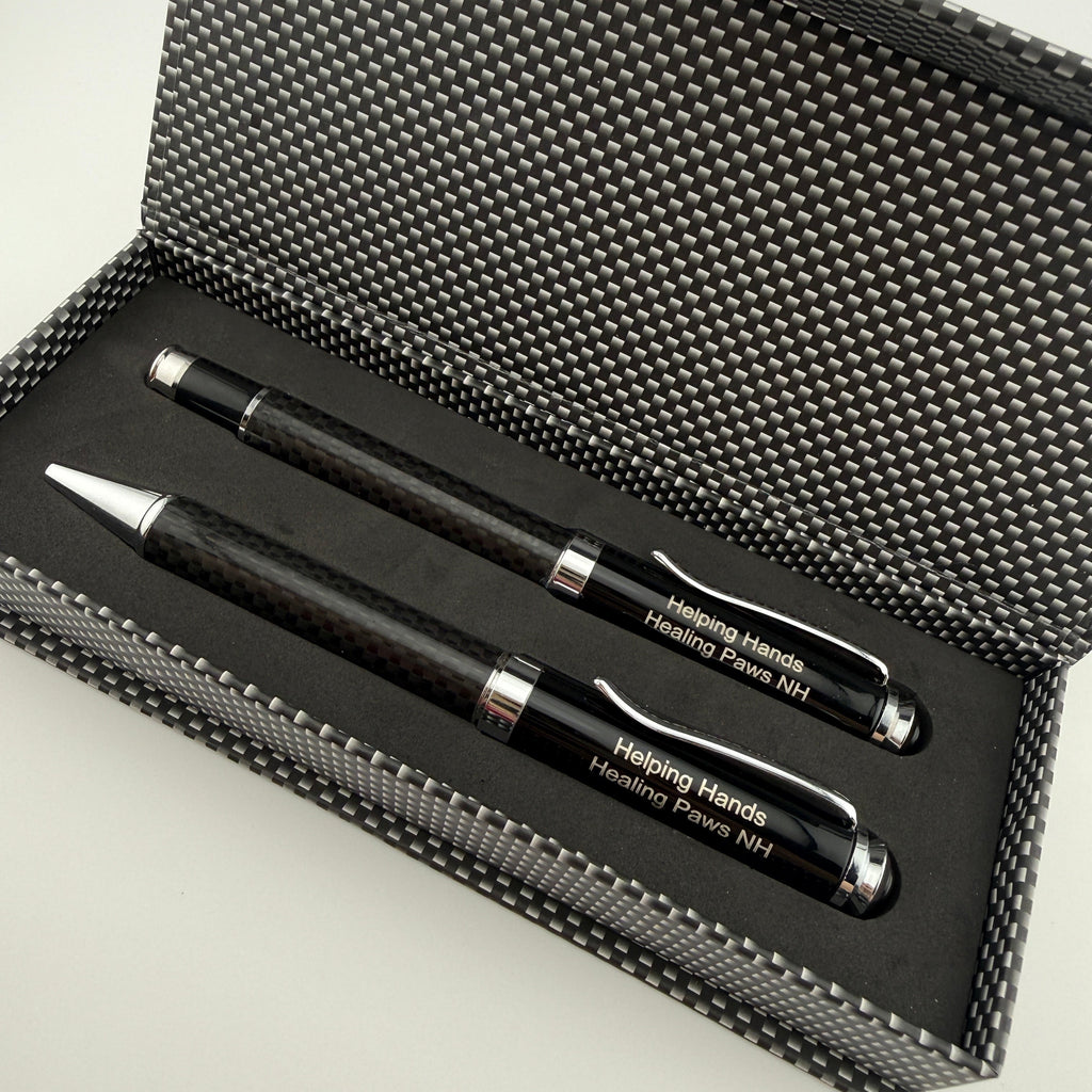 A black carbon-fiber-patterned gift box opened to reveal two matching pens with silver accents and white engraved text reading 'Helping Hands Housing Force N41'.