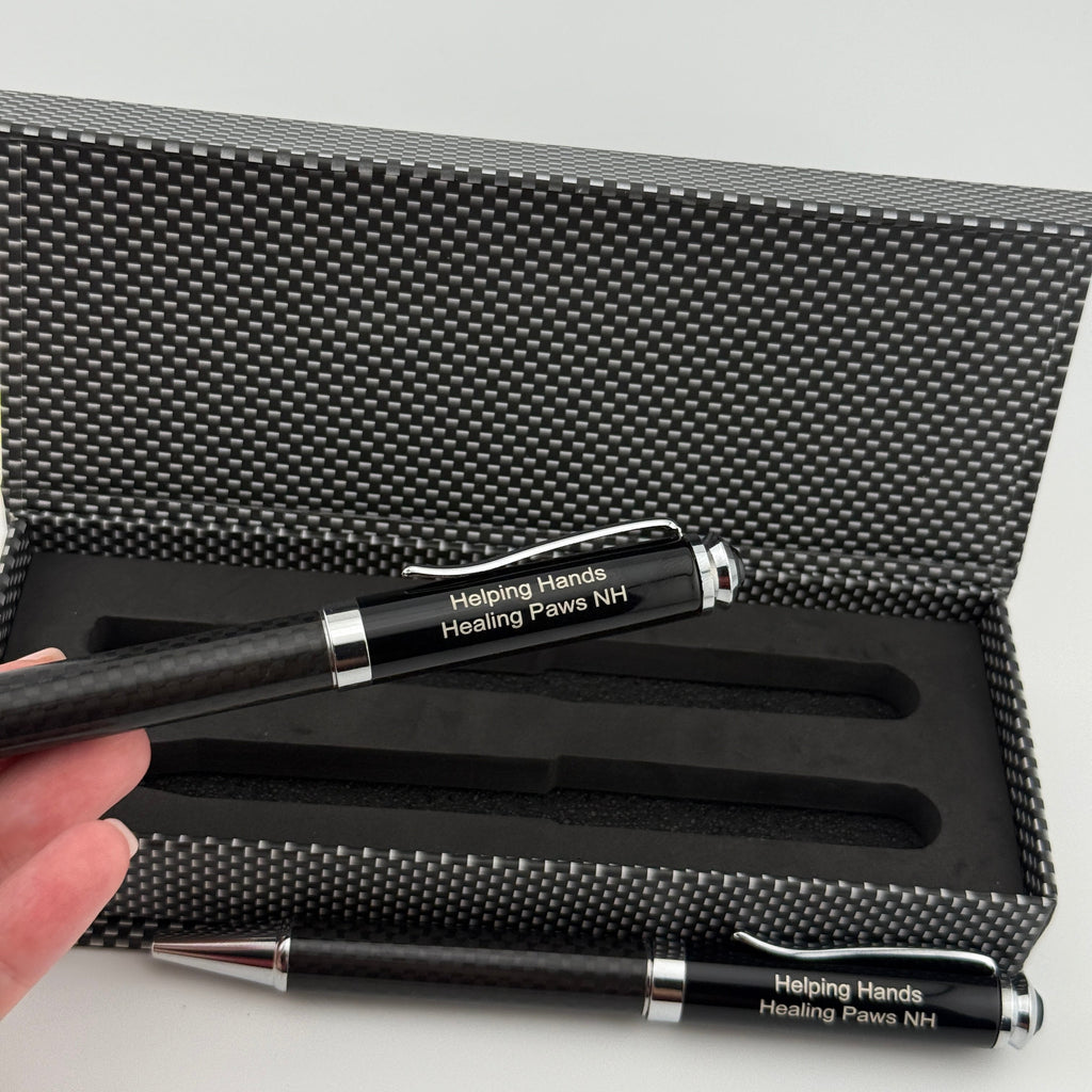 A black pen engraved with 'Helping Hands: Healing Paws NH' is held in a hand in front of an open textured black presentation box.