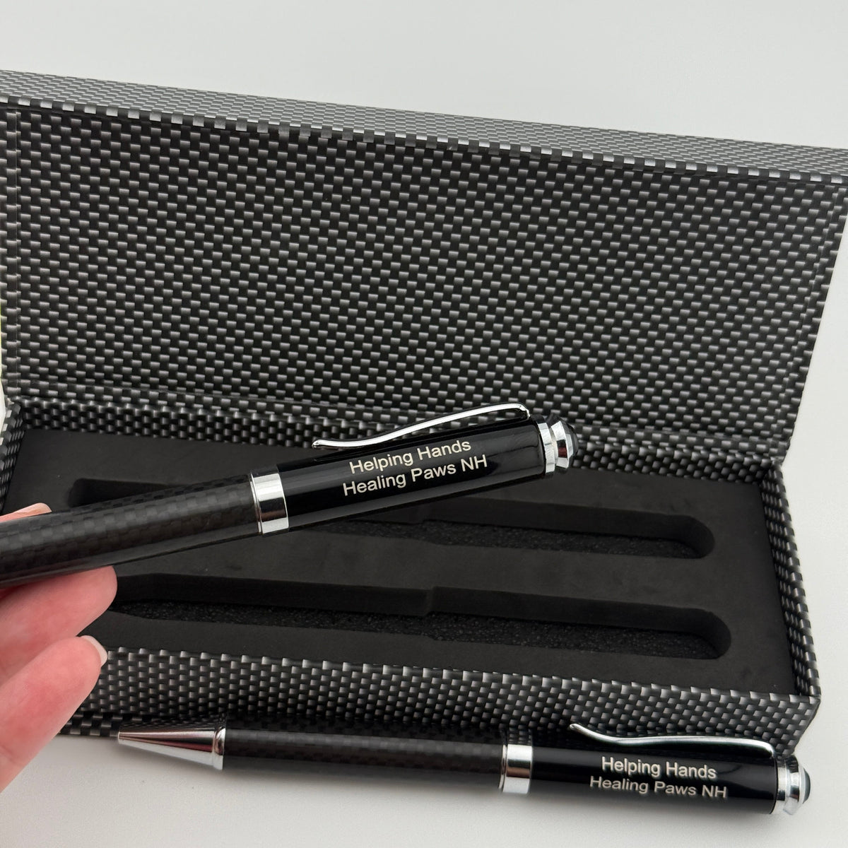 A black pen engraved with 'Helping Hands: Healing Paws NH' is held in a hand in front of an open textured black presentation box.