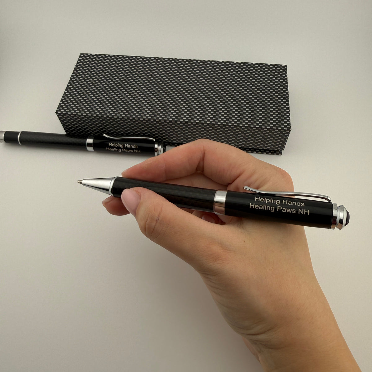 A hand holds a black ballpoint pen with silver trim and white printed text reading 'Helping Hands Healing Paws NH'. A second pen and a black textured box are visible in the background on a light neutral surface.