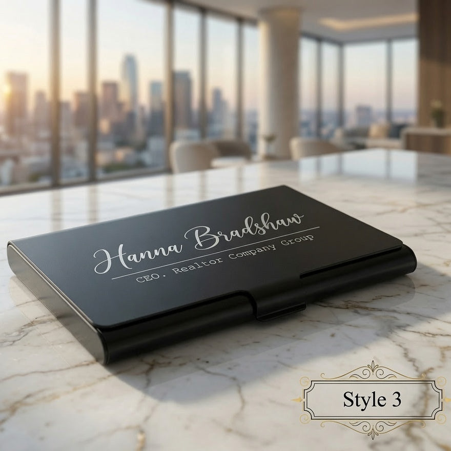 A black metal business card case with white script text reading 'Hanna Bradshaw' and smaller text 'CEO - Real Estate Company Group' sits on a white marble table in a modern office setting with a city skyline visible through large windows. A decorative label in the bottom right says 'Style 3'.