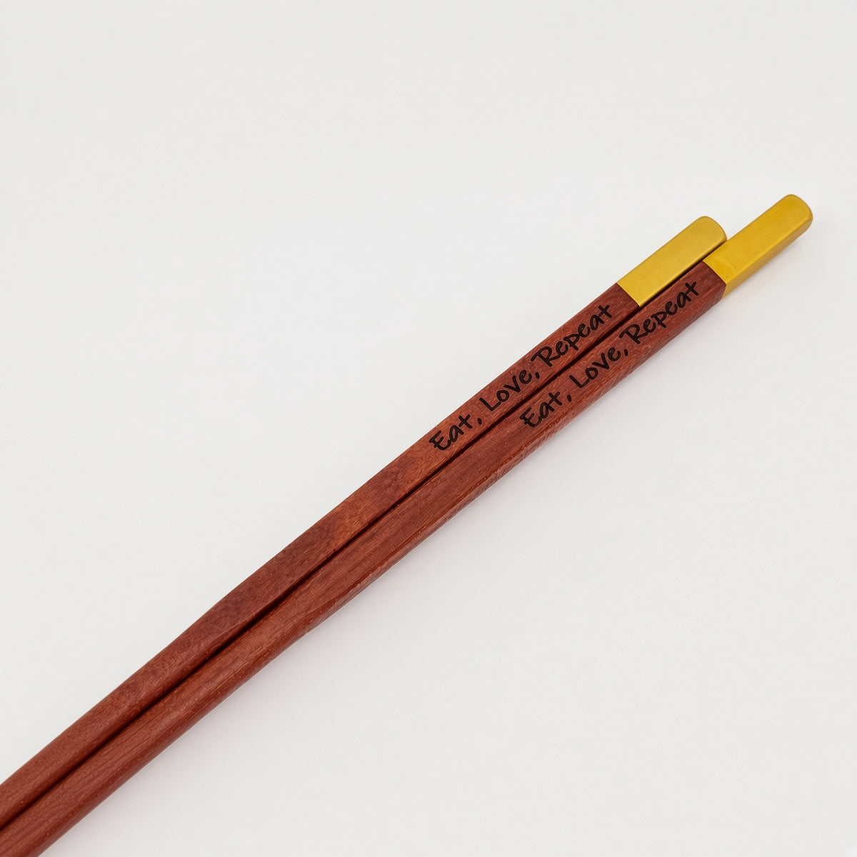 A pair of wooden chopsticks with a reddish-brown finish and bright gold-colored tips. The shafts are engraved with the words 'Eat, Love, Repeat' in black script.