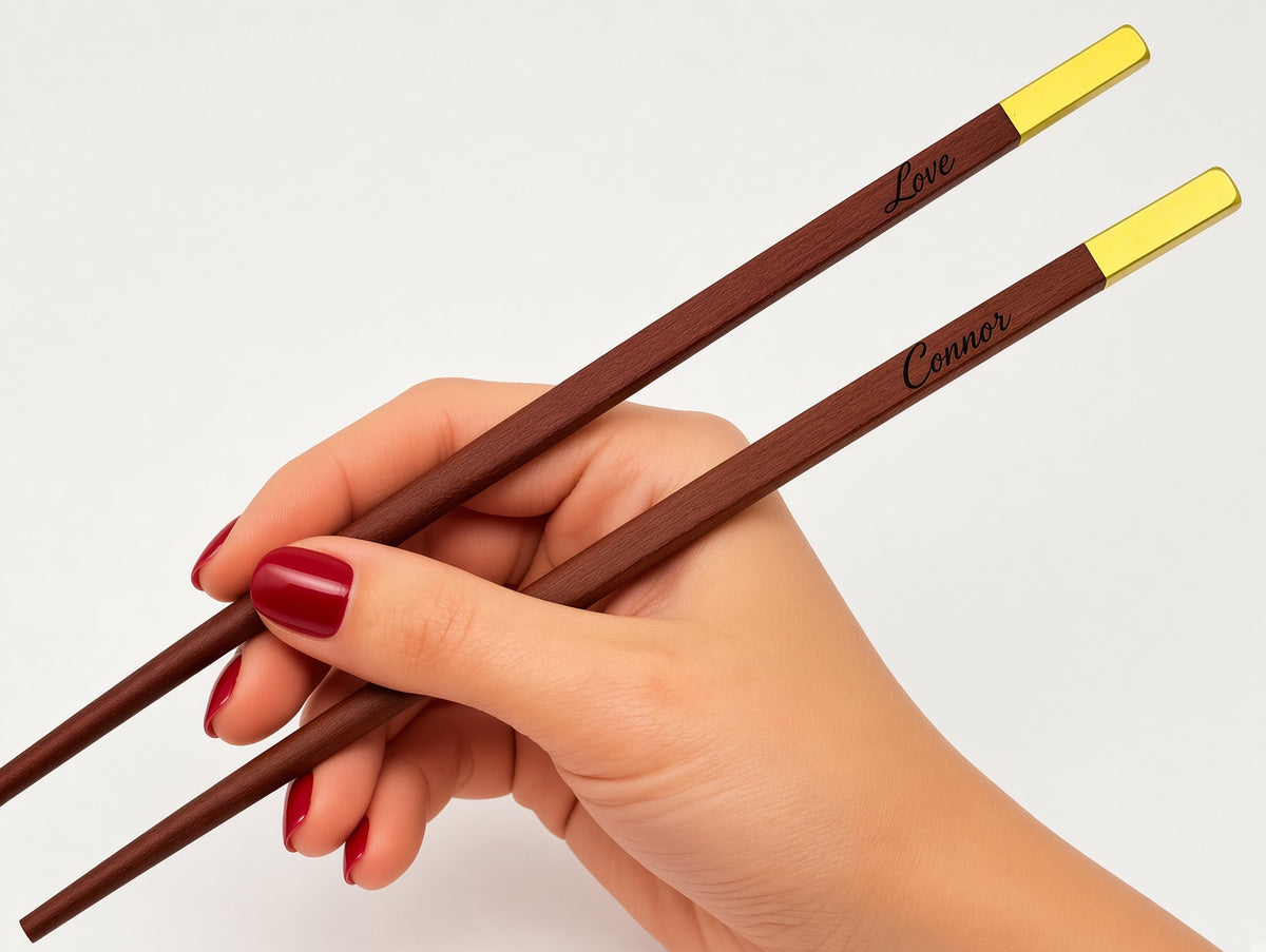 A hand with red nail polish holds a pair of dark brown wooden chopsticks with gold metal tips. The chopsticks are personalized with the names 'Love' and 'Connor' engraved near the gold ends, set against a plain white background.