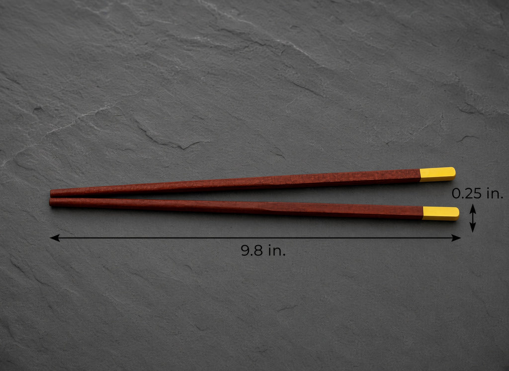 A pair of reddish-brown wooden chopsticks with bright yellow tips, displayed on a dark gray slate background with dimension lines indicating a length of 9.8 inches and a tip width of 0.