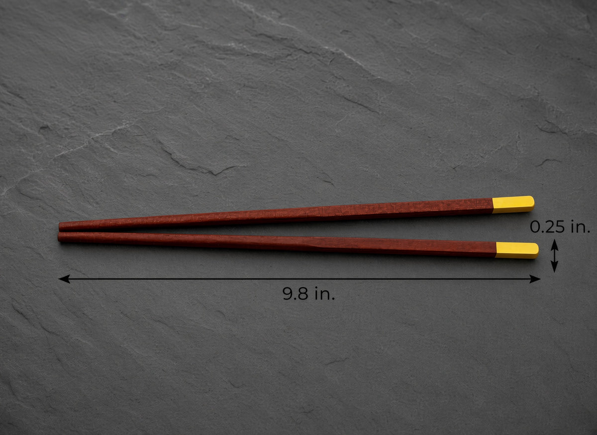 A pair of reddish-brown wooden chopsticks with bright yellow tips, displayed on a dark gray slate background with dimension lines indicating a length of 9.8 inches and a tip width of 0.