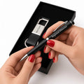 A black ballpoint pen engraved with 'Regional Director' and 'Alex Carter' is held by two hands with red nail polish, positioned in front of an open black gift box containing a matching black and silver keychain.