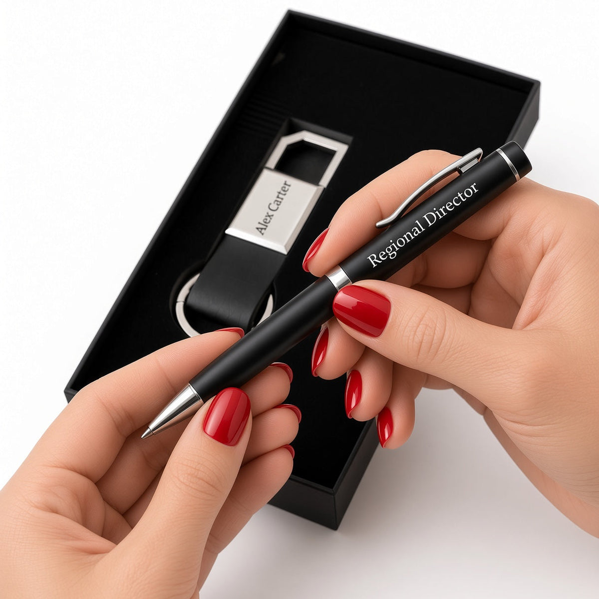 A black ballpoint pen engraved with 'Regional Director' and 'Alex Carter' is held by two hands with red nail polish, positioned in front of an open black gift box containing a matching black and silver keychain.