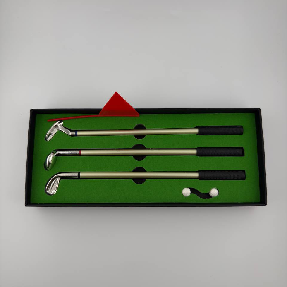 A set of three golf clubs with silver metal shafts and black grips, presented in a black gift box with a green interior. The set includes a red flag, two white golf balls, and a small black-and-white club head.