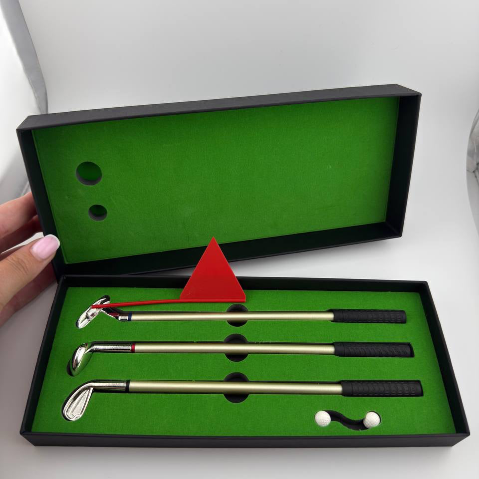A golf club set displayed in an open black presentation box with a bright green interior. The set includes three metallic golf clubs with black grips, a red triangular marker, and two white golf balls.